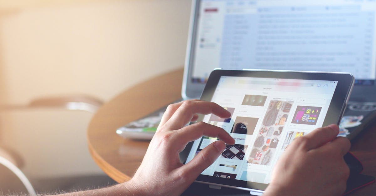 Hands using a tablet beside a laptop in a home office setting, showcasing online browsing.