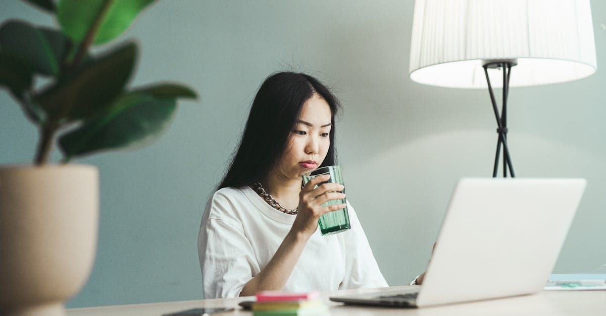 An Asian woman working at home on her laptop, drinking water.