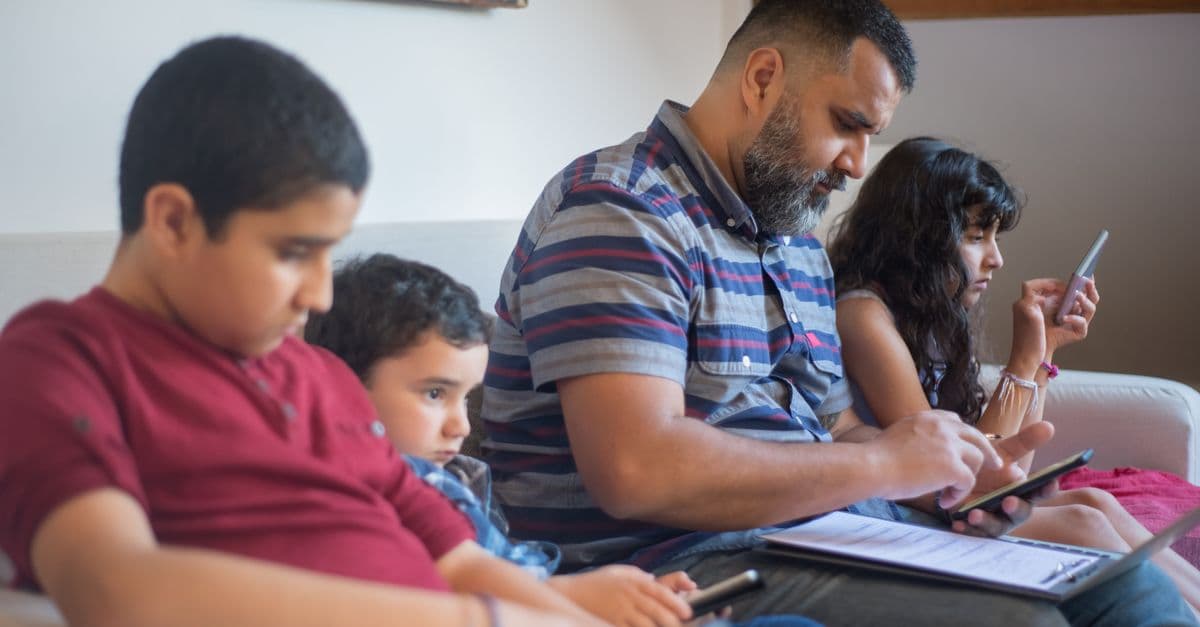 A father and three children engaging with technology while sitting on a sofa indoors.