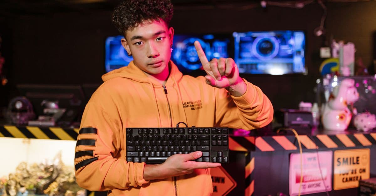 A young man in an orange jacket poses with a mechanical keyboard in a gaming setup.