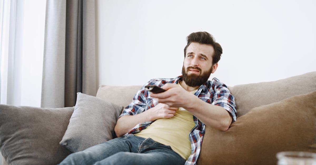 Casual bearded man lounging on a sofa, watching TV and relaxing in a cozy living room.