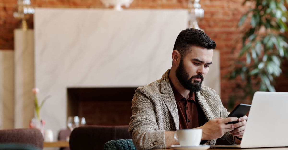 Bearded man using smartphone and laptop for remote work in a trendy coffee shop setting.
