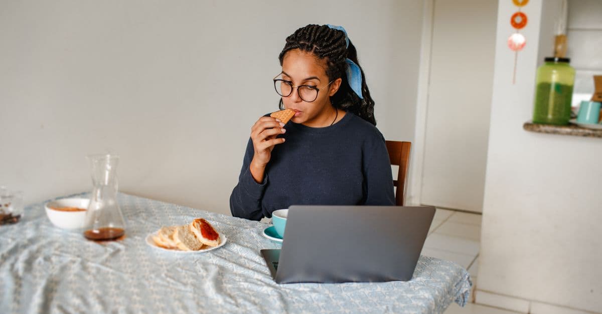 A woman eating breakfast and working on a laptop at a dining table indoors.