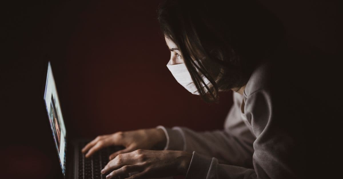A woman wearing a face mask focuses on her laptop screen in dim lighting, highlighting remote work.