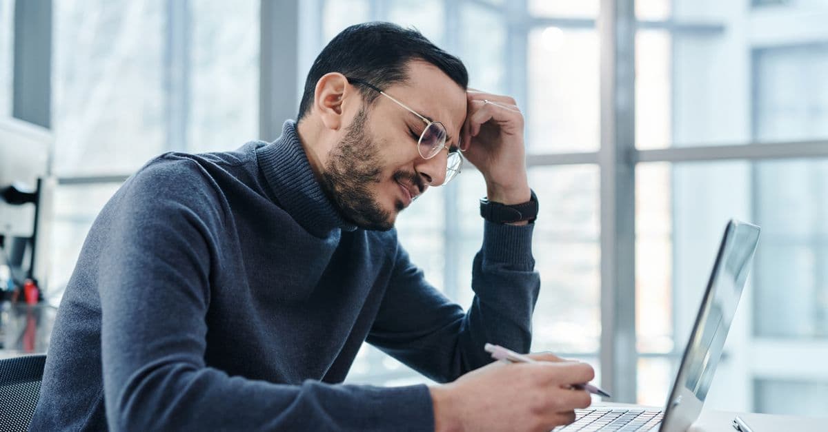Man looking stressed at laptop in a modern office with large windows.