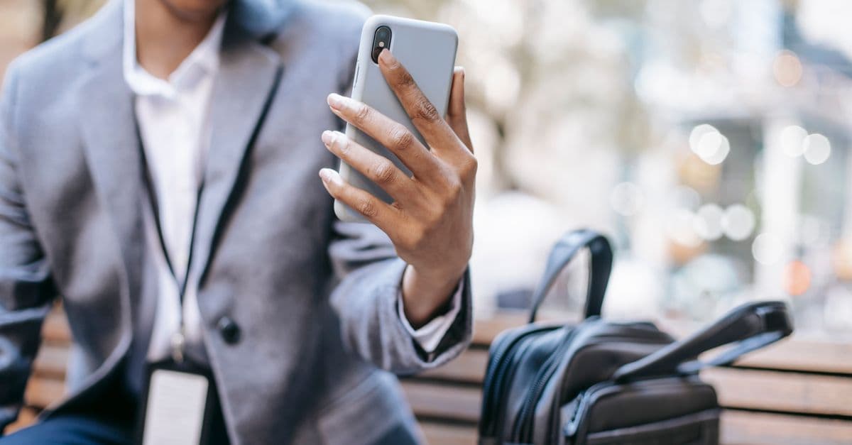 Crop unrecognizable happy young African American female employee in formal outfit and name tag smiling while having video call on mobile phone sitting on bench on city street