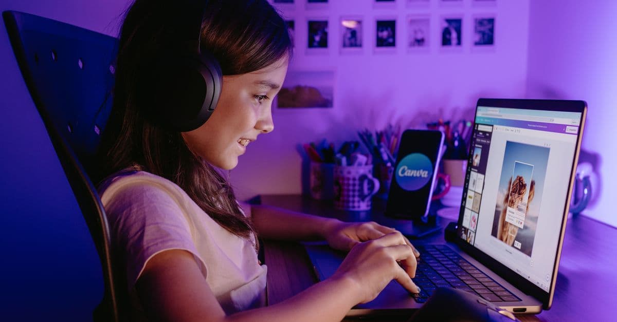 A young girl wearing headphones works on graphic design using a laptop and smartphone in a cozy, creative workspace.
