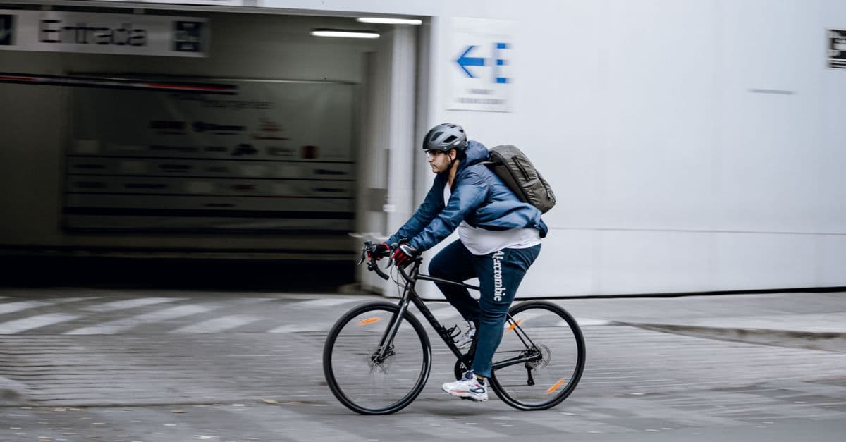 A man in a blue jacket cycling on an urban sidewalk with a backpack.