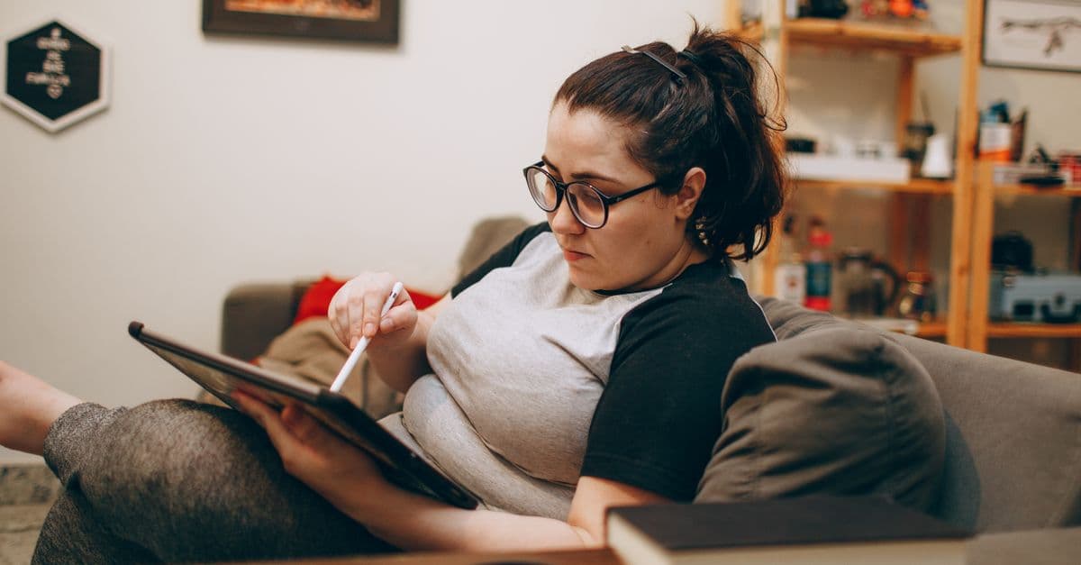 Woman wearing glasses, relaxing on couch while using tablet. Perfect for tech and lifestyle themes.