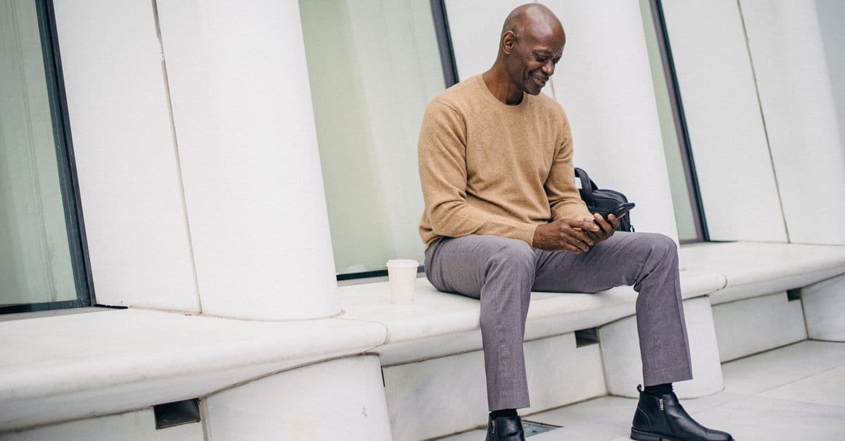 Full body of African American bald male in elegant clothes resting on stone bench texting on smartphone