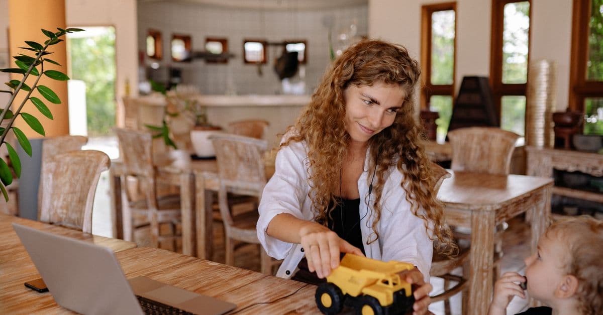 A mother with curly hair playing with her toddler son and a toy truck at a wooden table indoors.