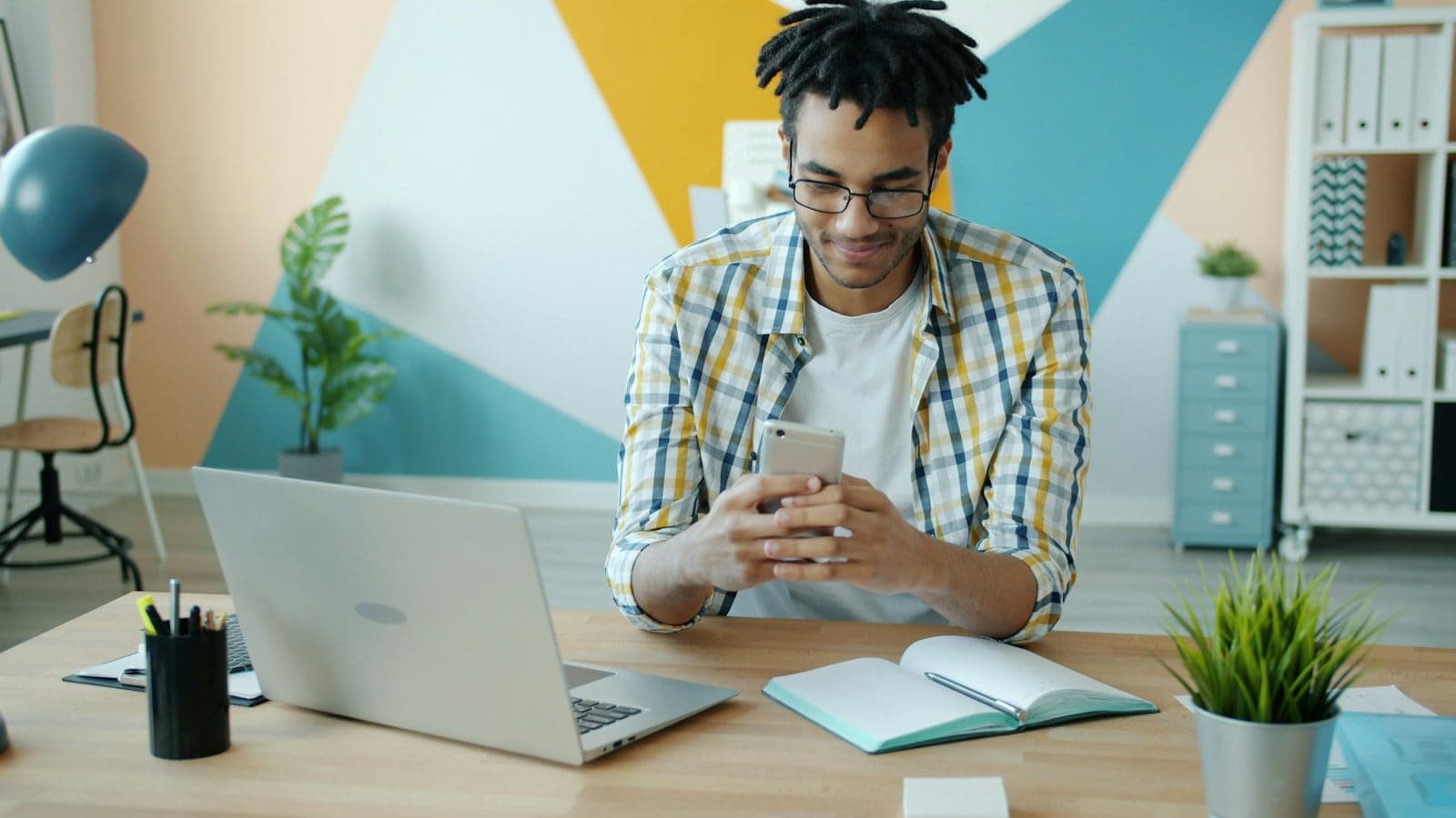 Man looking at phone at de k with laptop and plant