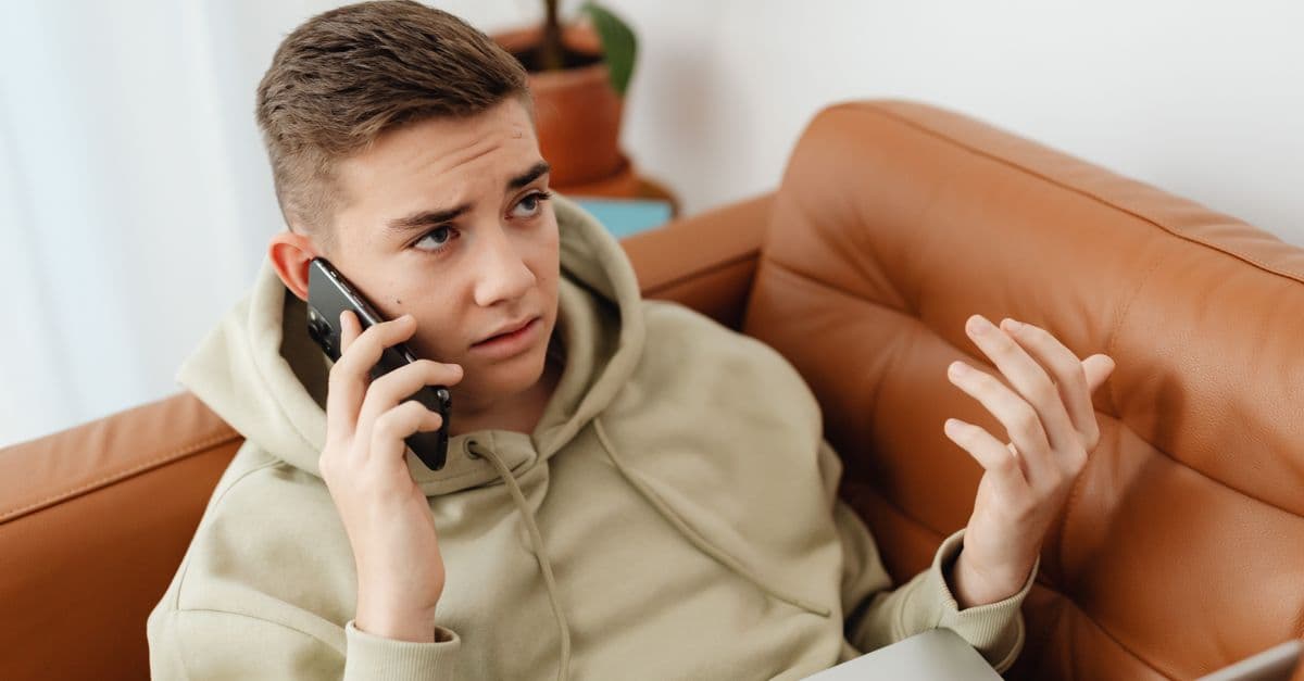 A young man talks on the phone while working on a laptop, looking concerned and frustrated.