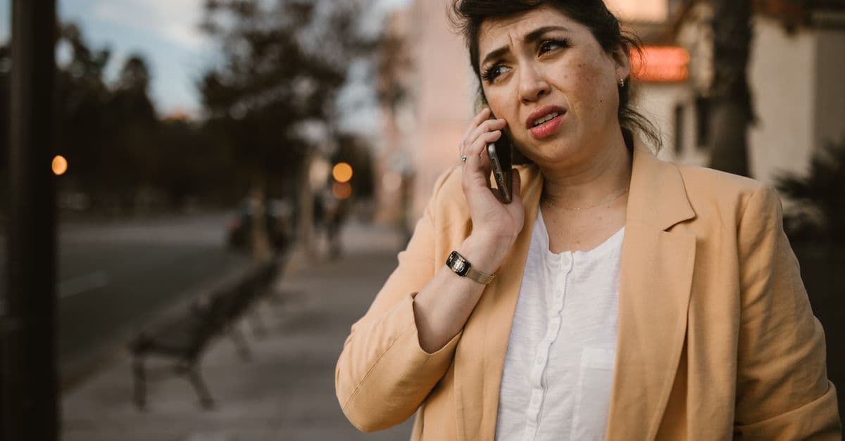 Woman in beige jacket looks concerned while making phone call outdoors.