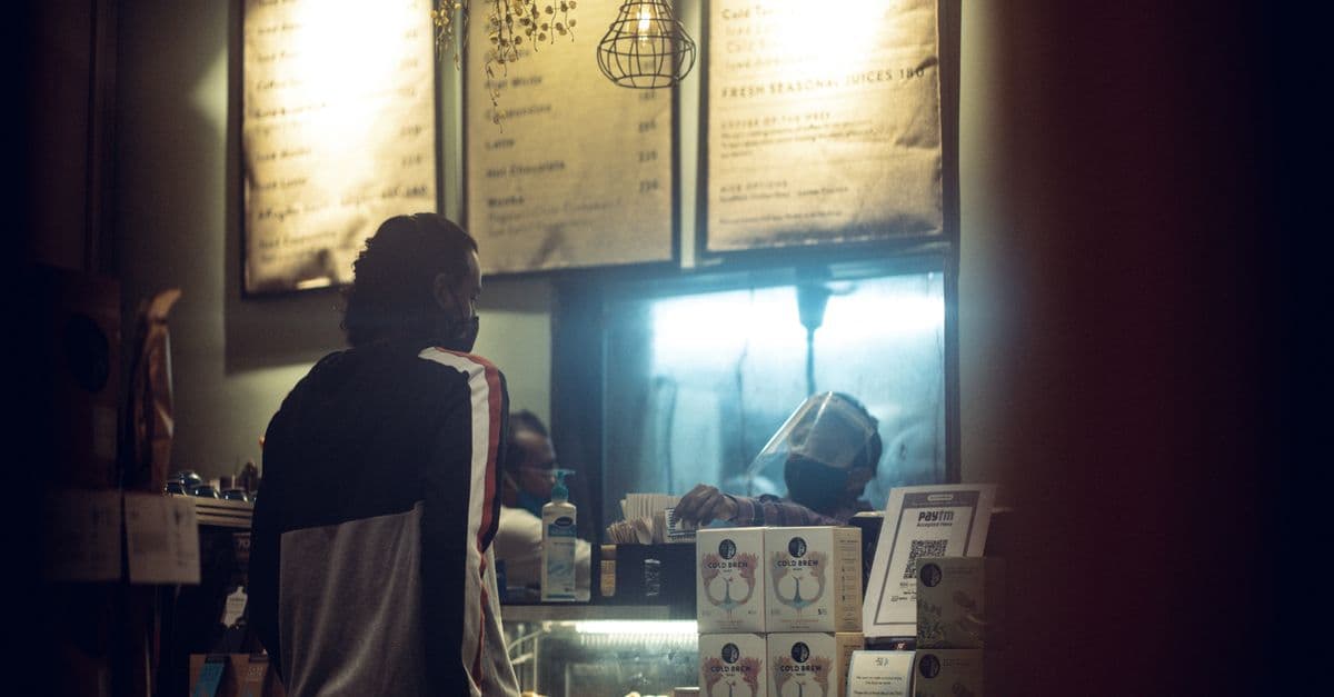 A customer wearing a mask orders at a warmly lit cafe counter during night time.