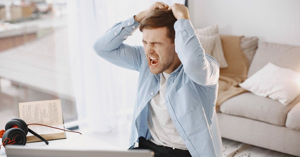 An upset man with mouth open, pulling hair at a desk with a laptop in a home setting.