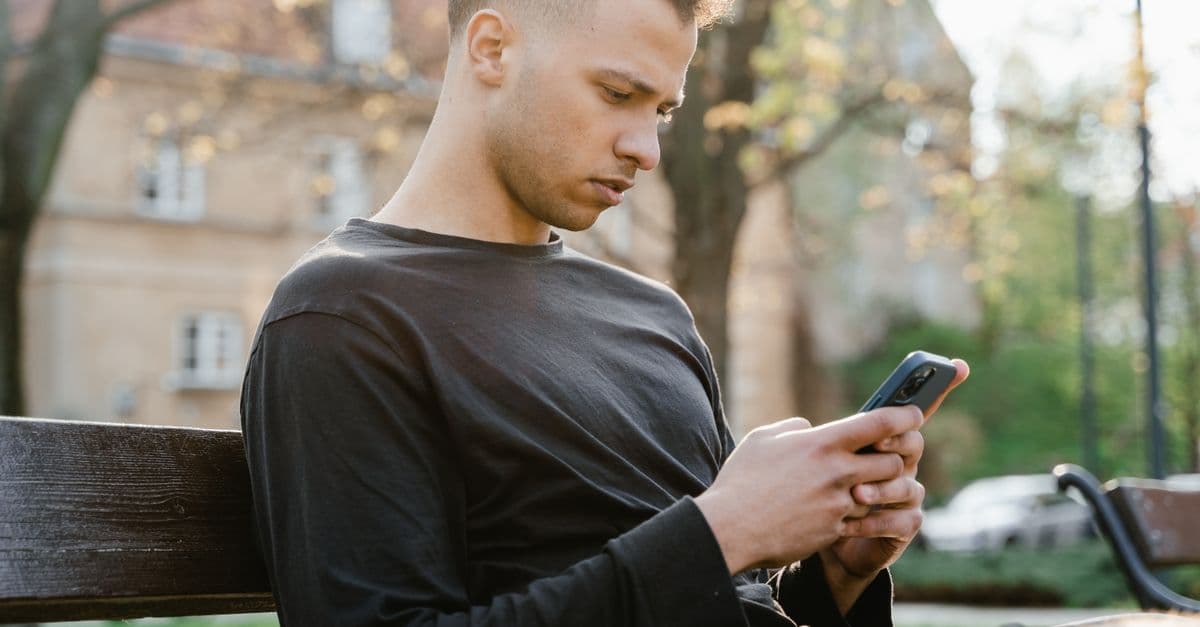 A young man sitting on a park bench using his smartphone during a sunny day.