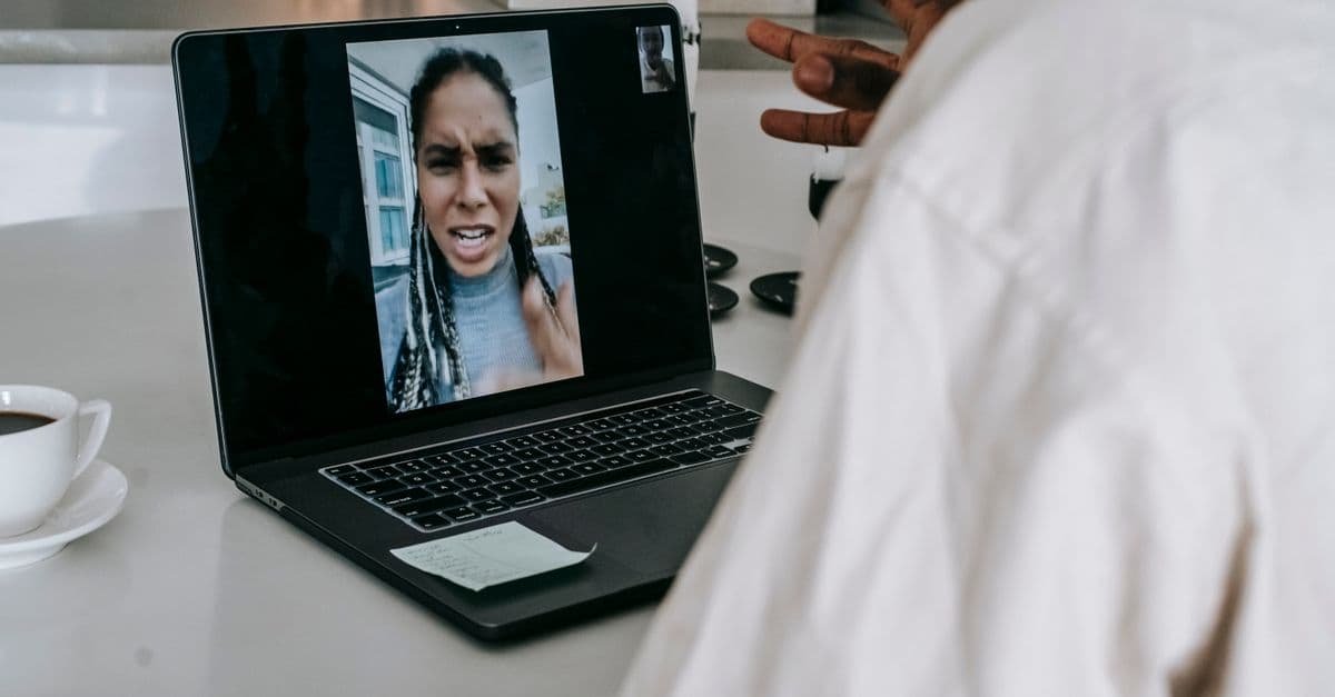 Crop anonymous black male sitting at table in modern kitchen with cup of coffee while having video conversation on laptop and arguing with angry girlfriend