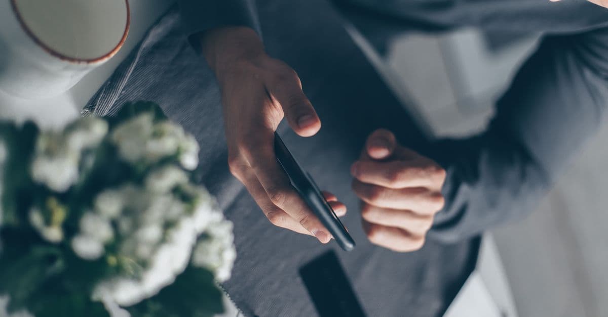 Top view of a man's hands holding a smartphone indoors with a cup and flowers nearby.