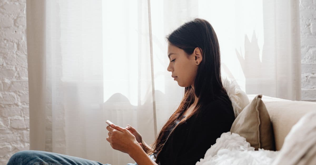 A teenager sits comfortably on a couch, focused on her smartphone in a peaceful indoor setting.