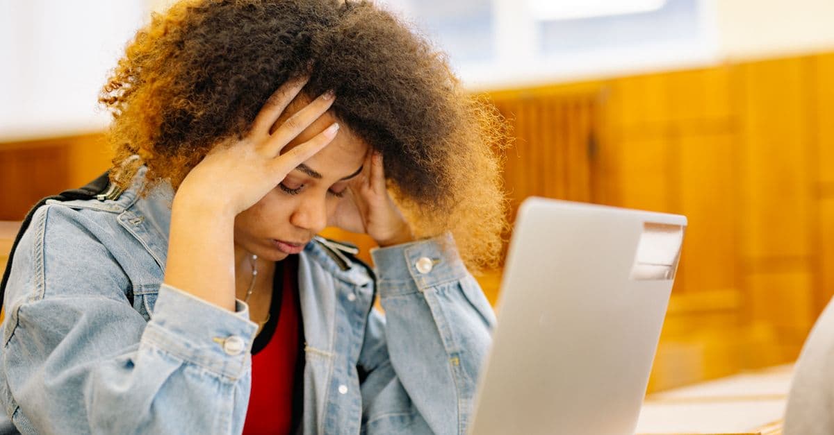 A young woman experiencing a headache while studying with a laptop indoors, looking stressed and overwhelmed.