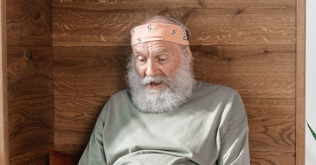 Elderly man with a beard shows surprise while using a laptop indoors.