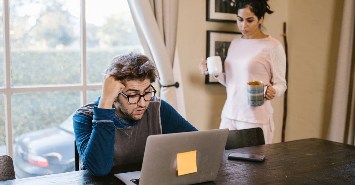 A man appears stressed working on a laptop at home while a woman brings him a drink.