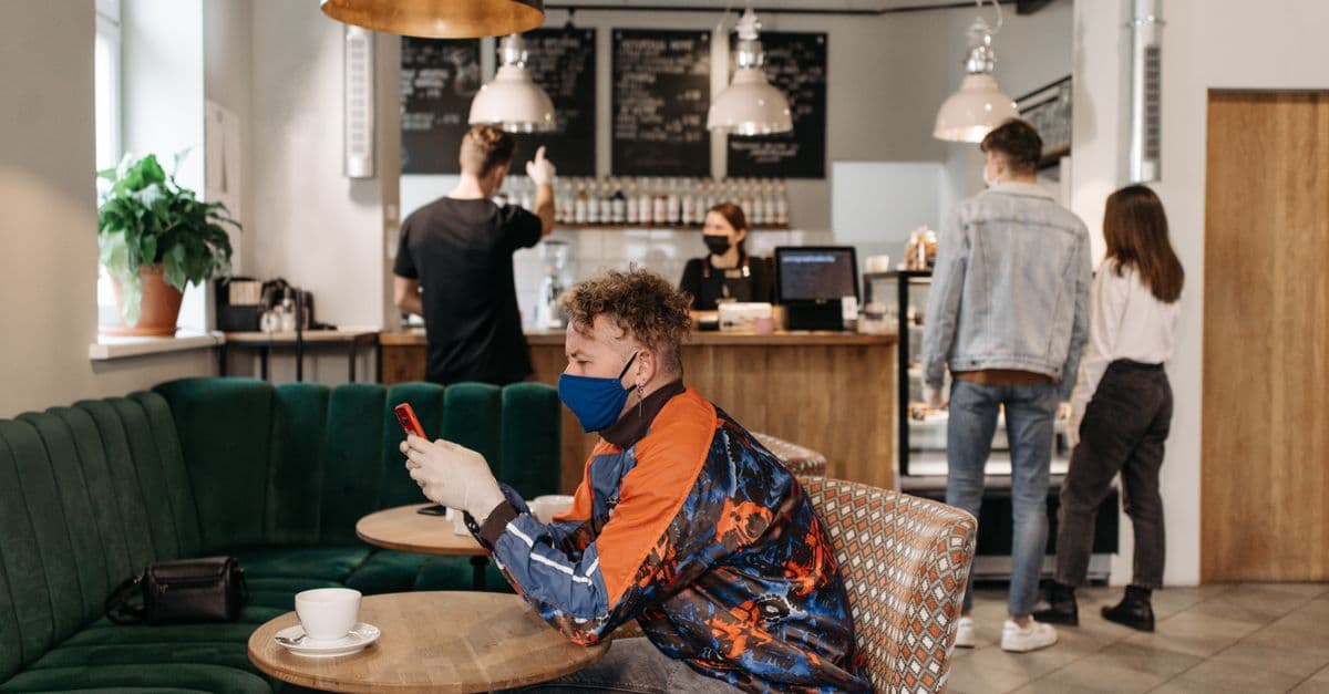A man wearing a face mask uses his phone in a stylish café with people around.