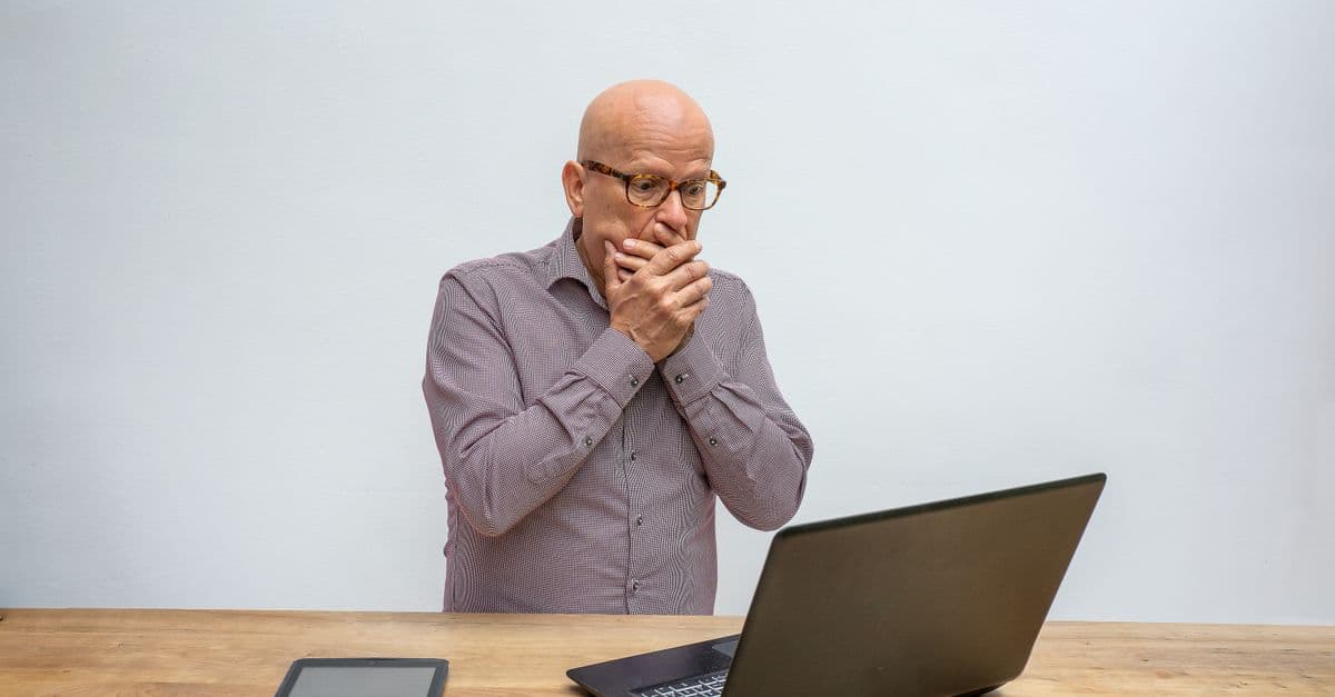 Elderly man reacts in surprise to laptop screen at wooden desk with tablet and mouse.
