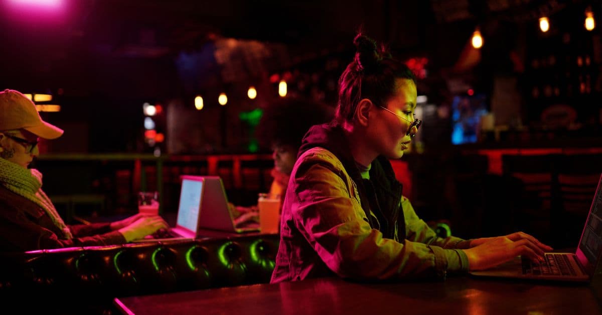 Asian woman typing on laptop in a cozy, dimly lit cafe with stylish illumination.