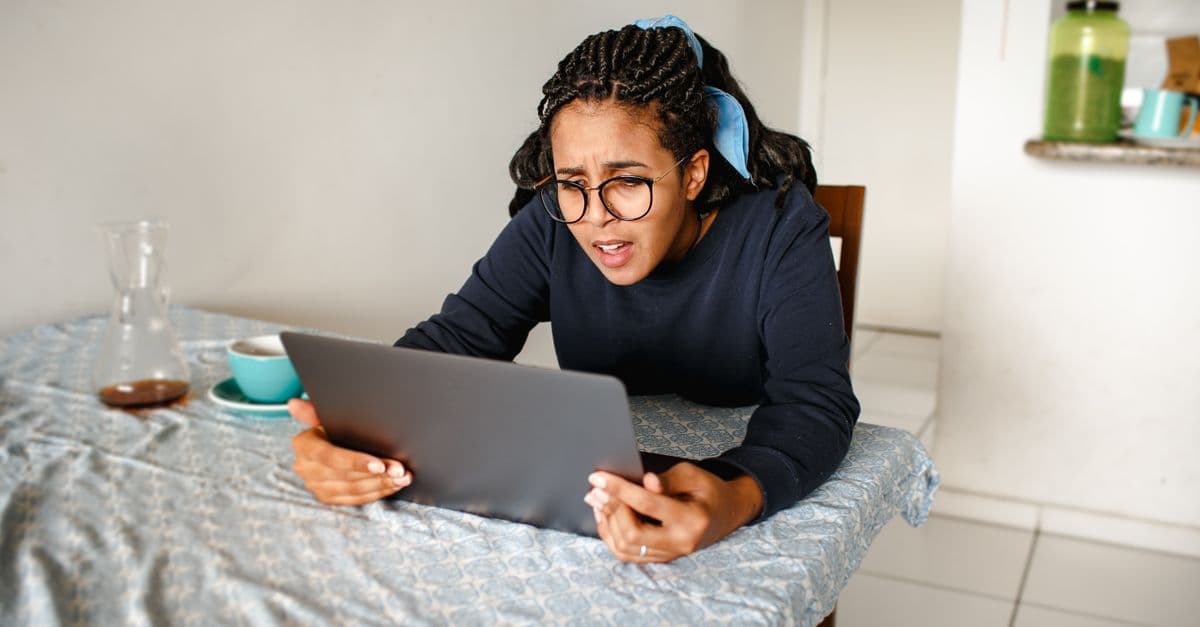 A woman with glasses intently focusing on her laptop screen at a dining table indoors.