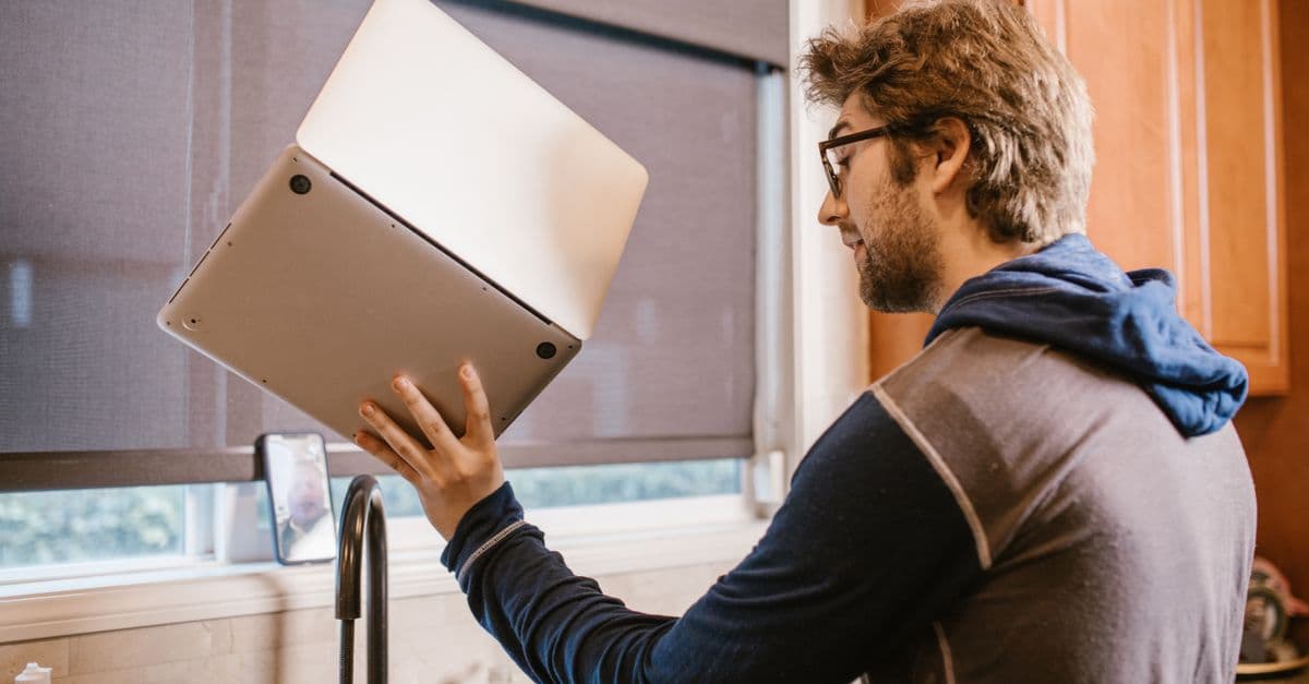 A man holding a laptop near a kitchen sink, examining it closely indoors.
