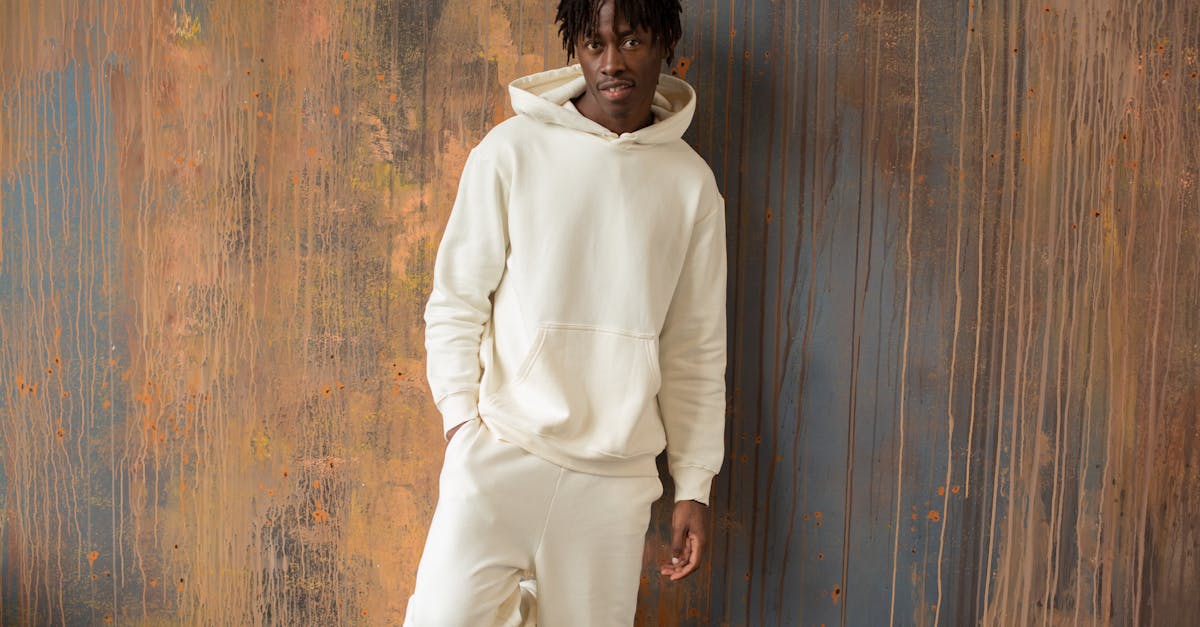 Content African American male in white hoodie and pants standing with hand in pocket near shabby wooden wall and looking at camera