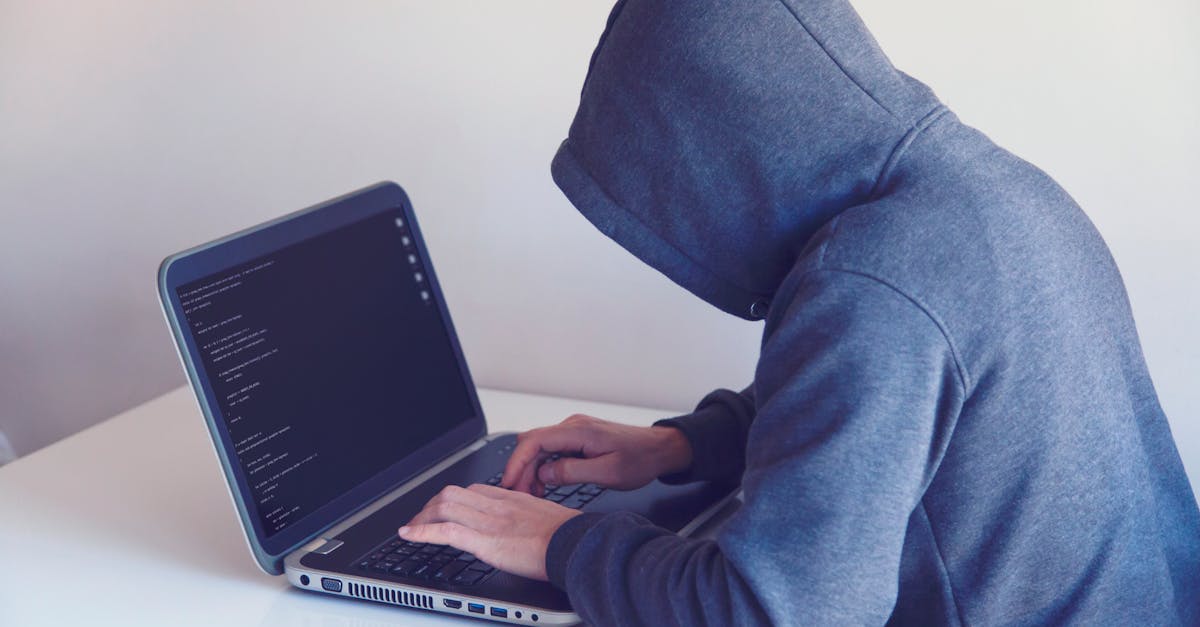 Side view of unrecognizable hacker in hoodie sitting at white table and working remotely on netbook in light room near wall
