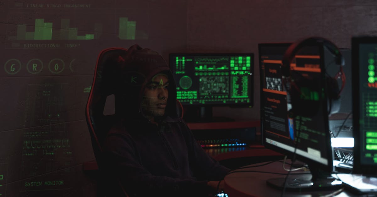 A man in a black hoodie engaged in cybersecurity work using multiple monitors indoors.
