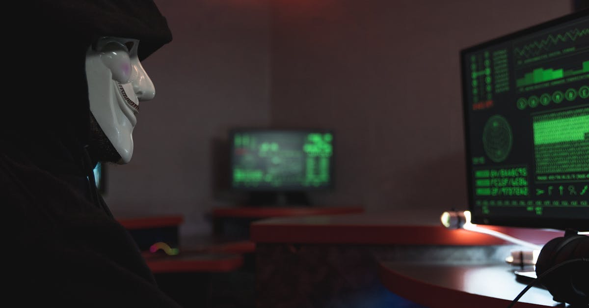 A person wearing a hacker mask operates a computer in a dimly lit room with digital displays.