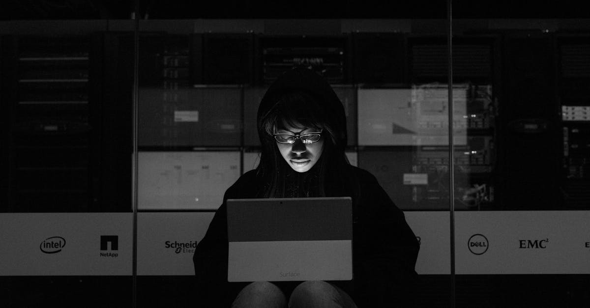 A woman in a dark room uses a laptop, illuminated by its screen, with server equipment in the background.