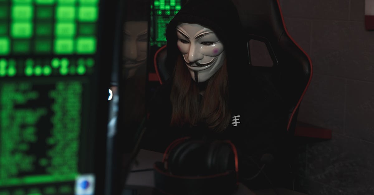 A mysterious hacker wearing a Guy Fawkes mask and black hoodie in a dimly lit room focused on computer screens.
