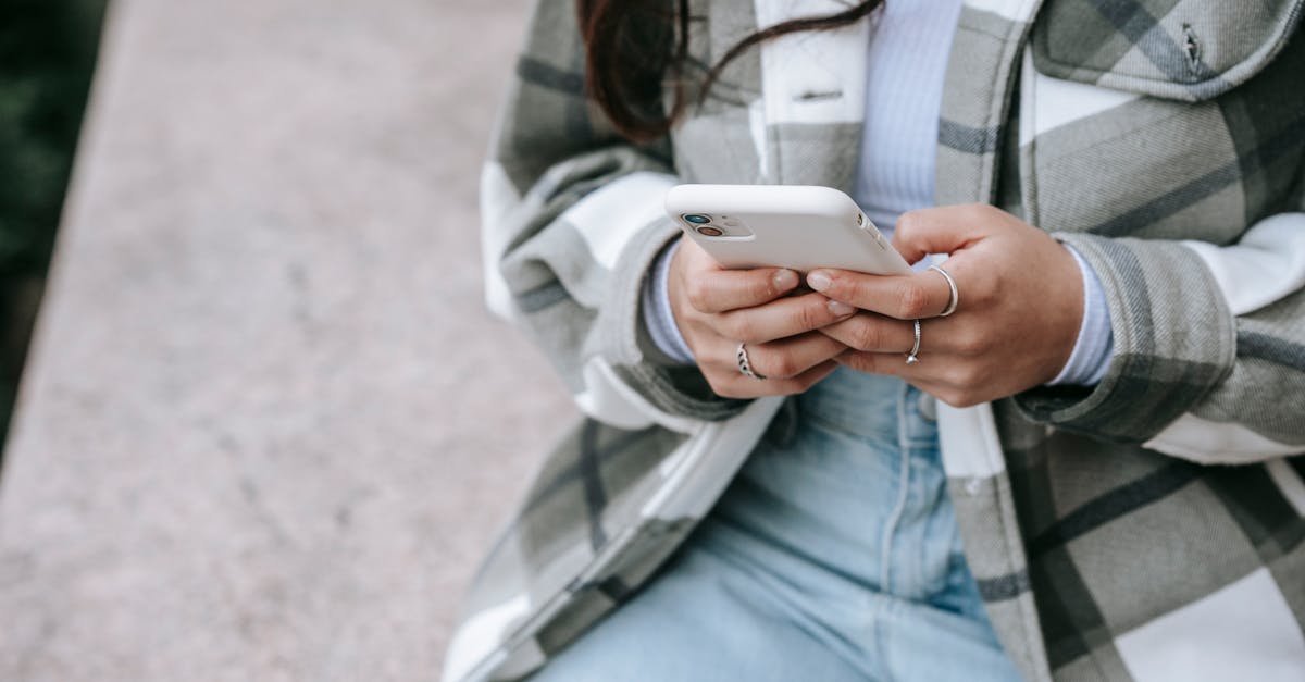 Crop unrecognizable young female in casual clothes sitting on stone surface and checking notifications on smartphone in daylight