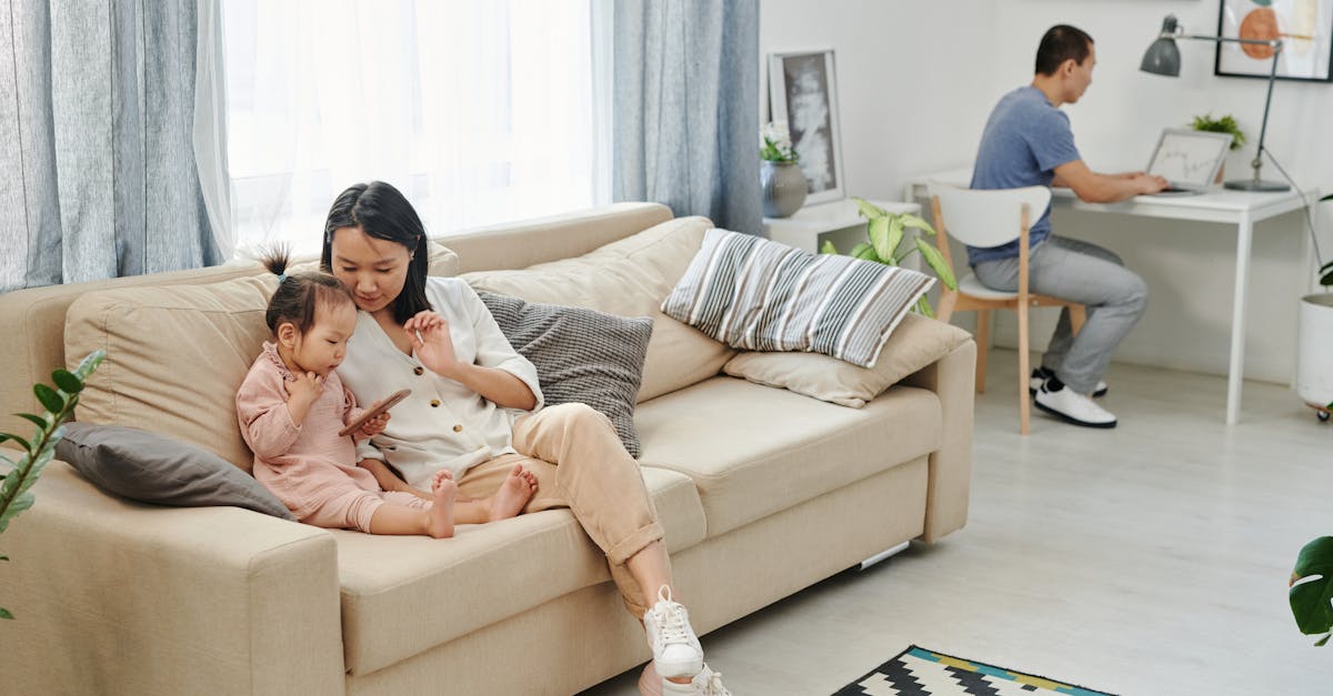 A mother and child bonding on a sofa while a man works on a laptop in a well-lit living room.