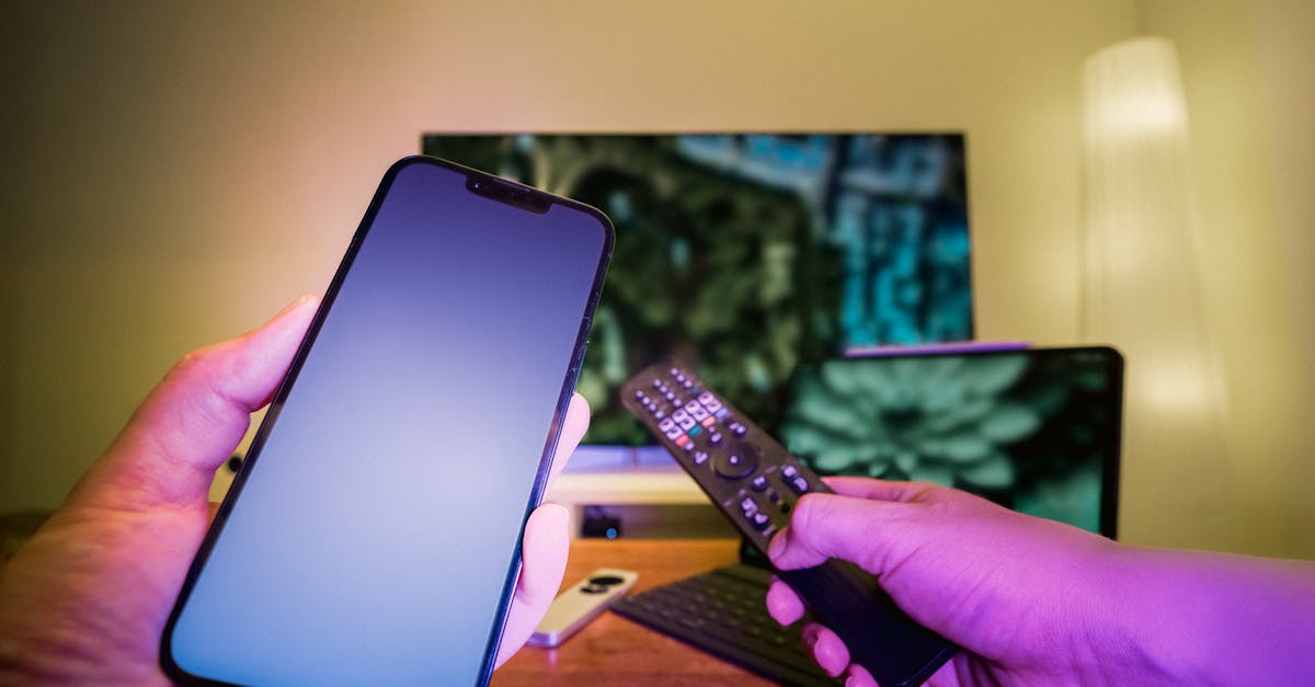 Hands holding a smartphone and remote control in a high-tech home setup with TV and other electronic devices.