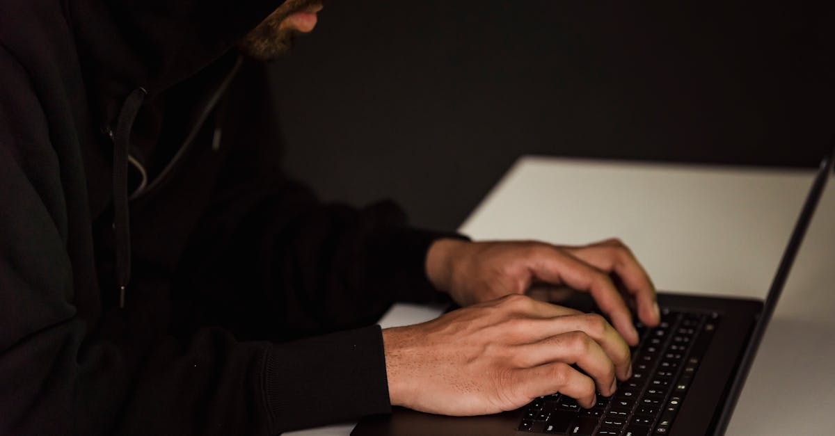 Crop bearded male hacker in black hood typing on netbook keyboard and browsing dark net in obscure room