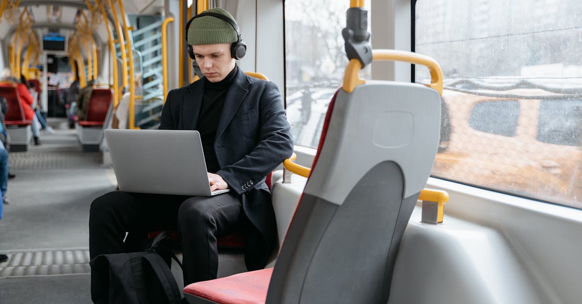 A man in a black coat using a laptop while commuting on a tram, listening to music.