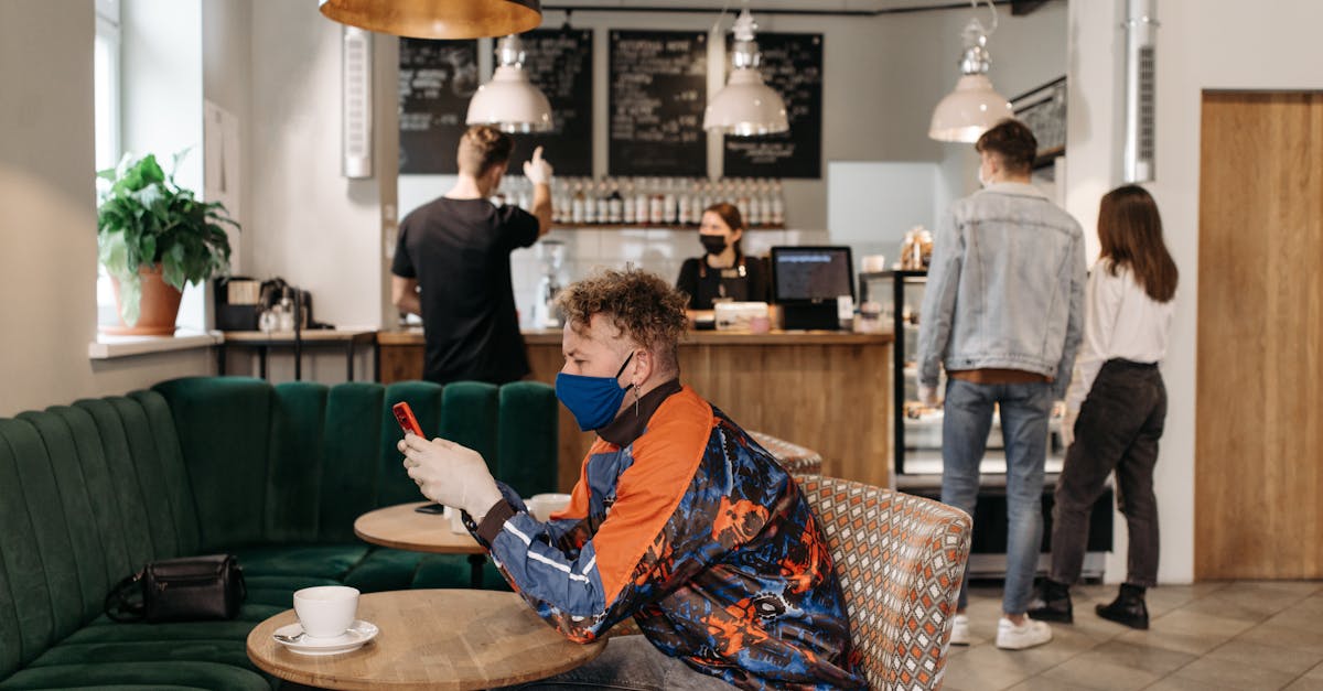 A man wearing a face mask uses his phone in a stylish café with people around.