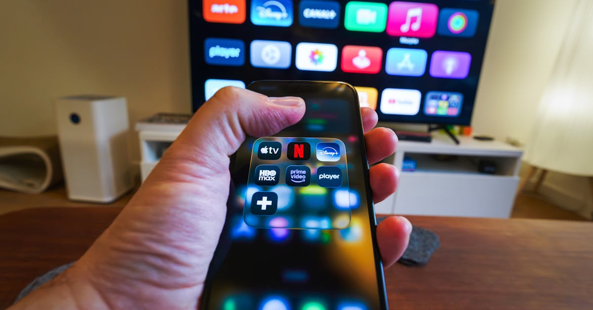 Close-up of a hand holding a phone displaying streaming apps in front of a TV with multiple app icons.