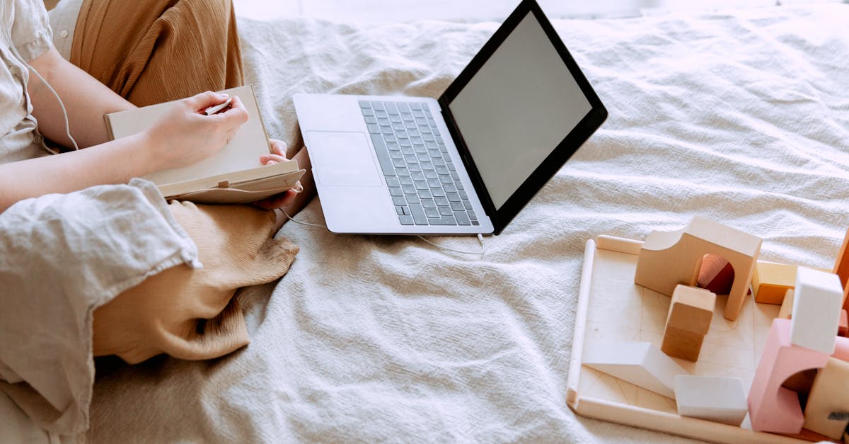 Unrecognizable female using laptop and writing notes in notepad while sitting on bed and working on project in bedroom on sunny day