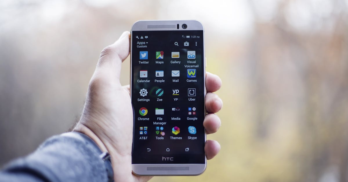 A close-up of a hand holding an HTC smartphone with visible apps on the screen, in an outdoor setting.
